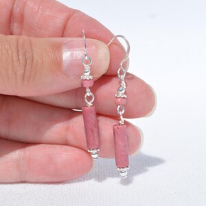 May include: A pair of drop earrings featuring cylindrical pink stone beads suspended from silver-toned metal hooks. Each earring includes a small round pink bead accent above the cylindrical stone, connected by silver-toned metal links.