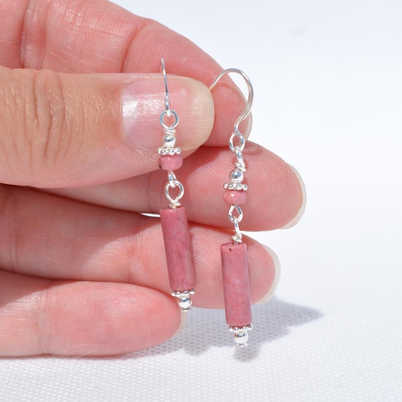 May include: A pair of drop earrings featuring cylindrical pink stone beads suspended from silver-toned metal hooks. Each earring includes a small round pink bead accent above the cylindrical stone, connected by silver-toned metal links.