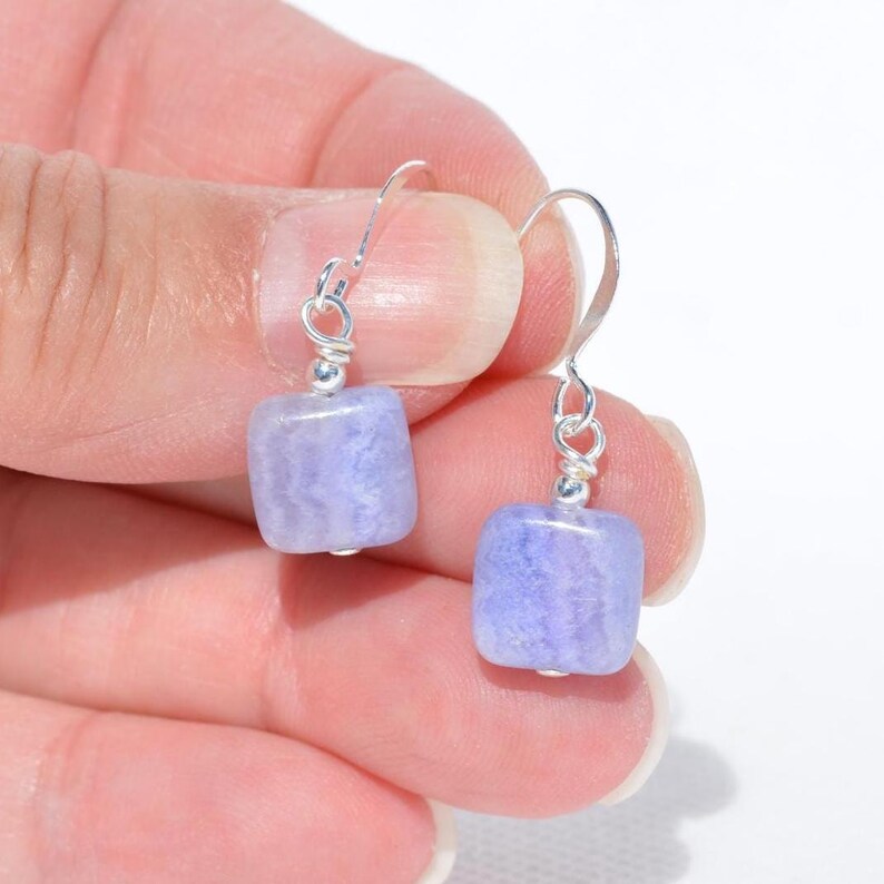 May include: A pair of earrings featuring light purple, square-shaped beads. The beads are suspended from silver-colored ear wires. The earrings are held up against a white background.