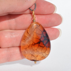 May include: A teardrop-shaped pendant made of orange agate with brown and black markings. The pendant is attached to a gold chain.