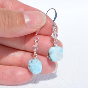 May include: A pair of dangle earrings featuring square, light blue gemstones with dark flecks. The earrings have silver-colored hooks and are accented with clear, round beads. The earrings are held up by a hand.