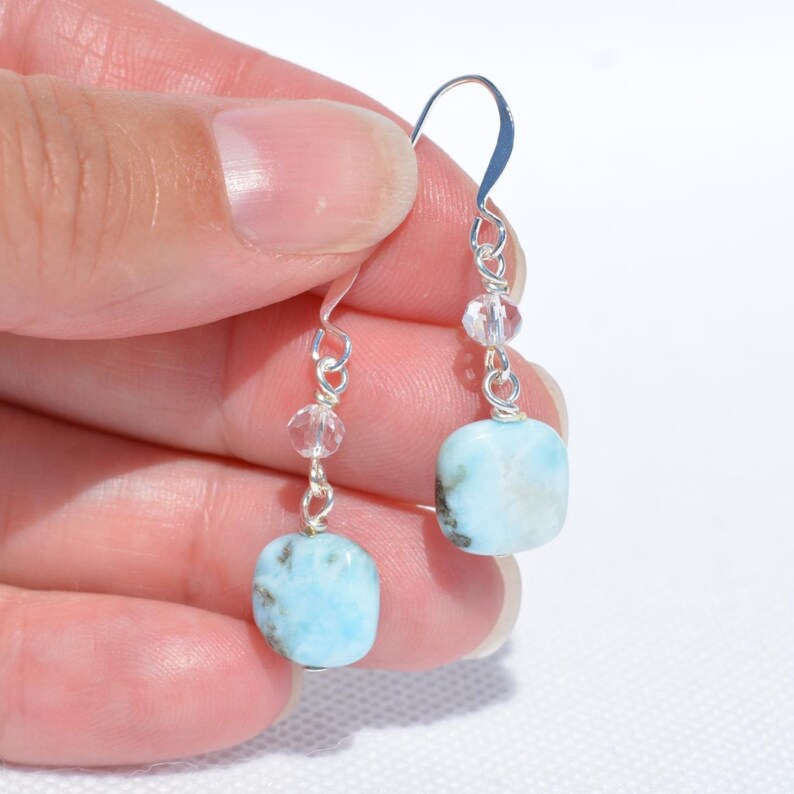 May include: A pair of dangle earrings featuring square, light blue gemstones with dark flecks. The earrings have silver-colored hooks and are accented with clear, round beads. The earrings are held up by a hand.