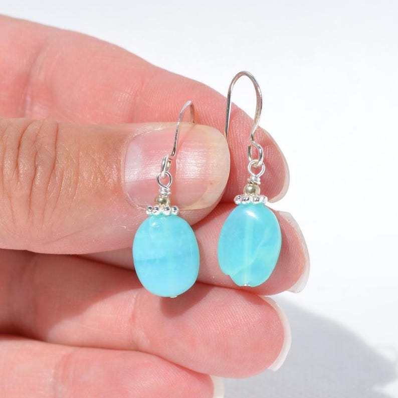 May include: A pair of drop earrings featuring smooth, oval-shaped light blue stones. Each stone is suspended from a silver-toned metal hook by a small silver wire wrap and a tiny silver bead, creating a simple and clean design for these earrings.