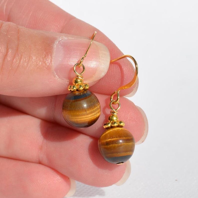 Puede incluir: Un par de pendientes colgantes con piedras preciosas de ojo de tigre redondas y pulidas con bandas marrones y doradas. Cada piedra cuelga de un gancho de metal dorado mediante un alambre dorado y un grupo de tres peque&ntilde;as cuentas met&aacute;licas.