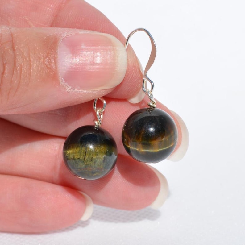 Op de afbeelding: Een paar hangende oorbellen met bolvormige tijgeroog-edelstenen met een chatoyante goudbruine en donkerblauwe glans. Elke gepolijste steen hangt aan een eenvoudige zilverkleurige metalen haak en heeft een glad, reflecterend oppervlak.