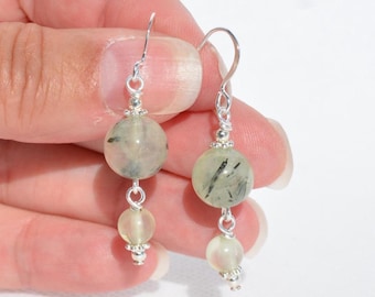 May include: A pair of dangle earrings featuring two round, pale green rutilated quartz beads with visible black needle-like inclusions. The beads are connected by silver-toned metal wire and small silver beads, hanging from silver-toned ear hooks.