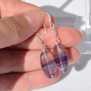 Puede incluir: Un par de pendientes colgantes con piedras ovaladas de fluorita translúcida púrpura y transparente. Los pendientes tienen ganchos plateados y pequeñas cuentas redondas. Las piedras miden aproximadamente 2,5 cm de largo.