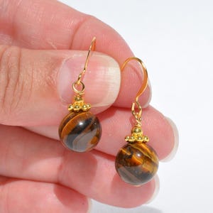 Puede incluir: Un par de pendientes colgantes con piedras preciosas de ojo de tigre redondas y pulidas con patrones de bandas marrones y doradas. Cada piedra est&aacute; rematada con una peque&ntilde;a tapa decorativa de metal dorado y cuelga de un gancho dorado.
