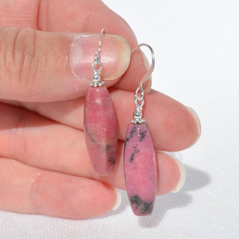 May include: A pair of drop earrings featuring elongated, cylindrical rhodonite beads in shades of dusty pink with dark grey veining. The stones are suspended from silver-toned metal hooks with small decorative silver beads at the top of each stone.