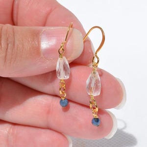 May include: A pair of gold-colored earrings. Each earring features a clear, teardrop-shaped stone, a gold chain, and a small, round, blue bead. The earrings are held up by a gold hook.