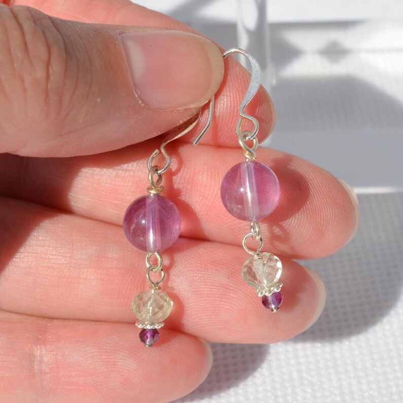 May include: A pair of dangle earrings featuring a large, round, translucent pink bead at the top, connected to a smaller, clear bead and a tiny, dark pink bead. The earrings have silver-colored hooks and connecting wires.
