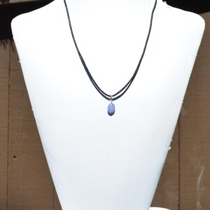 May include: A delicate necklace featuring a small, oval-shaped, light purple gemstone pendant. The pendant hangs from a thin, black cord necklace. The necklace is displayed on a white mannequin bust against a neutral background.