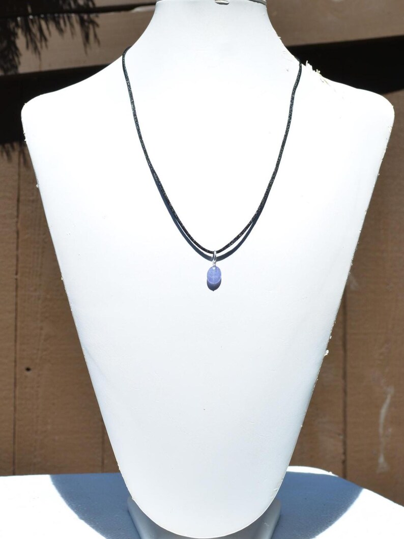 May include: A delicate necklace featuring a small, oval-shaped, light purple gemstone pendant. The pendant hangs from a thin, black cord necklace. The necklace is displayed on a white mannequin bust against a neutral background.