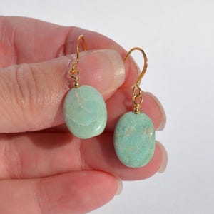May include: A pair of drop earrings featuring smooth, oval-shaped light green stones suspended from gold-toned metal hooks. Each stone displays natural veining and a polished finish, attached to the wire hook by a small gold-coloured metal bead.