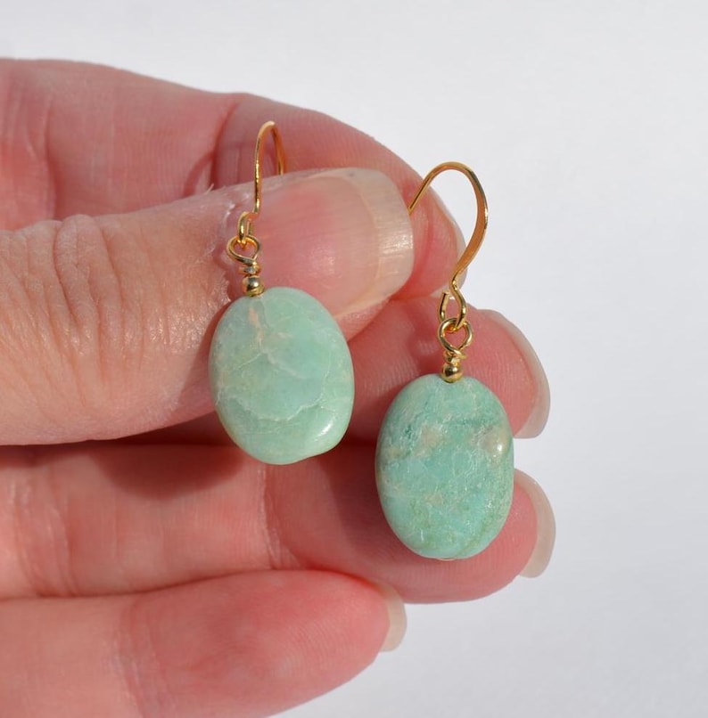 May include: A pair of drop earrings featuring smooth, oval-shaped light green stones suspended from gold-toned metal hooks. Each stone displays natural veining and a polished finish, attached to the wire hook by a small gold-coloured metal bead.