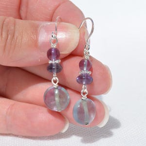 May include: A pair of drop earrings with silver hooks. Each earring has a large, round, translucent purple and blue bead at the base, connected to smaller purple beads and a silver accent. The earrings are shown against a light background.