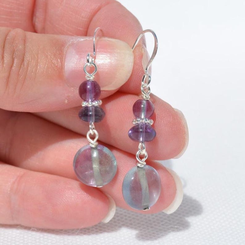 May include: A pair of drop earrings with silver hooks. Each earring has a large, round, translucent purple and blue bead at the base, connected to smaller purple beads and a silver accent. The earrings are shown against a light background.