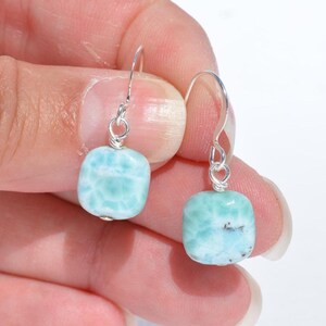 May include: A pair of earrings featuring square, light blue larimar stones with silver-colored hooks. The stones have a marbled appearance with white and darker blue inclusions, creating a unique, ocean-like aesthetic. The earrings are held by a person.