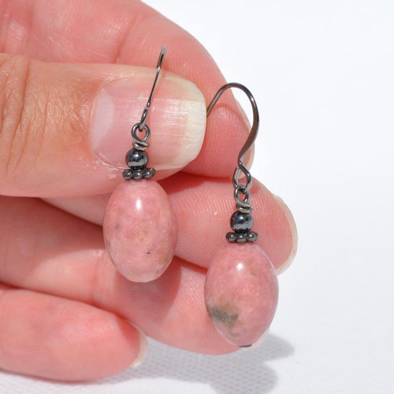 Peut inclure: Une paire de boucles d'oreilles pendantes compos&eacute;es de pierres de rhodonite rose ovales et lisses suspendues &agrave; des crochets en m&eacute;tal gris fonc&eacute;. Chaque boucle pr&eacute;sente une petite perle m&eacute;tallique sombre au-dessus de la pierre polie.