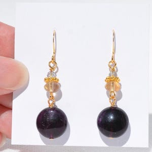 May include: Dangle earrings with gold-coloured hooks. Each earring has a clear bead, a cluster of small gold beads, a light orange bead, and a large, dark purple, round bead. The earrings are presented against a white background.