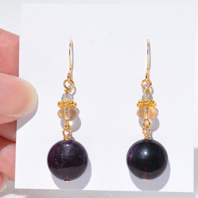 May include: Dangle earrings with gold-coloured hooks. Each earring has a clear bead, a cluster of small gold beads, a light orange bead, and a large, dark purple, round bead. The earrings are presented against a white background.