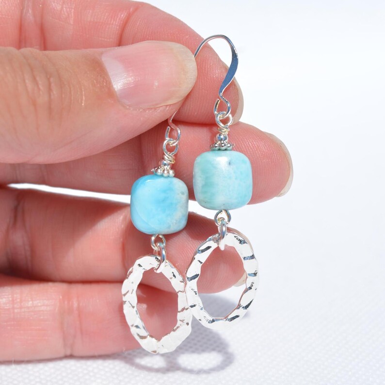 May include: A pair of dangle earrings featuring light blue, square-shaped beads and textured silver oval charms. The earrings are held up by silver hooks and are designed for pierced ears. The earrings are a handmade jewelry item.