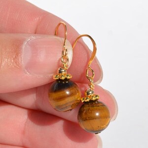Puede incluir: Un par de pendientes colgantes con piedras preciosas esf&eacute;ricas de ojo de tigre con bandas marrones y doradas. Cada piedra tiene una peque&ntilde;a tapa de metal dorado texturizado y cuelga de un gancho curvo de color dorado para orejas perforadas.