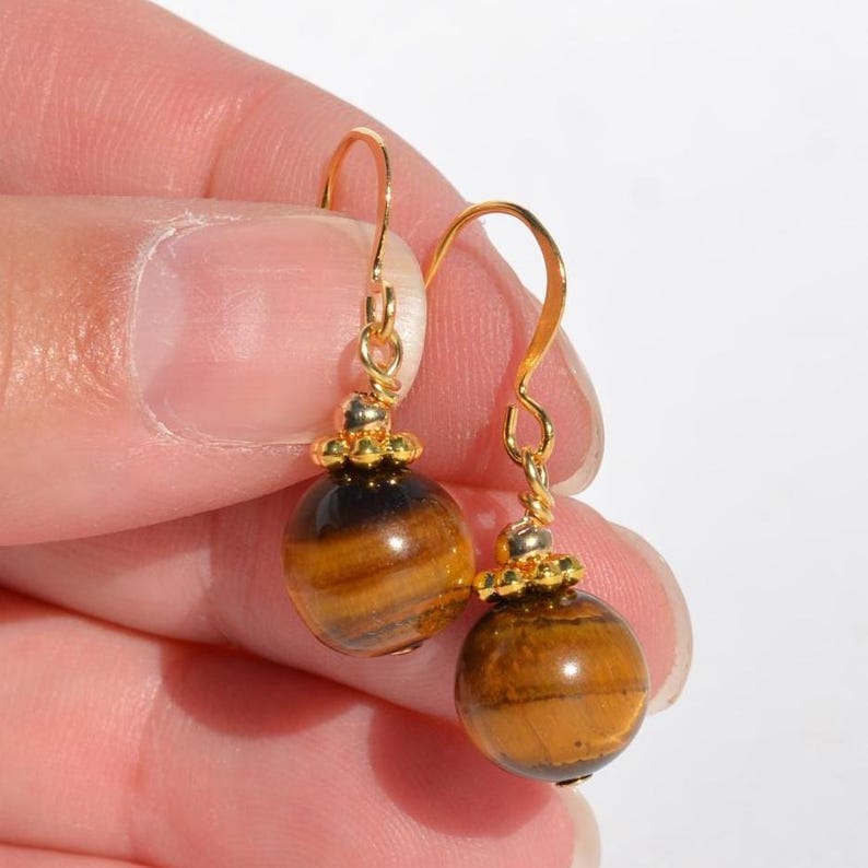Puede incluir: Un par de pendientes colgantes con piedras preciosas esf&eacute;ricas de ojo de tigre con bandas marrones y doradas. Cada piedra tiene una peque&ntilde;a tapa de metal dorado texturizado y cuelga de un gancho curvo de color dorado para orejas perforadas.