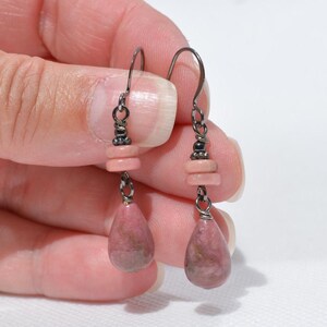 Peut inclure: Une paire de boucles d'oreilles pendantes avec des crochets en m&eacute;tal fonc&eacute; et des attaches en fil m&eacute;tallique. Chaque boucle comporte deux petites perles circulaires rose clair plates au-dessus d'un pendentif en pierre rose et gris.