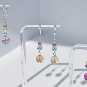 May include: Earrings with a silver hook and a combination of colorful beads. The earrings feature a large, round, purple and white striped bead at the bottom, with smaller beads in shades of pink, green, and yellow above. The earrings are displayed on clear stands.
