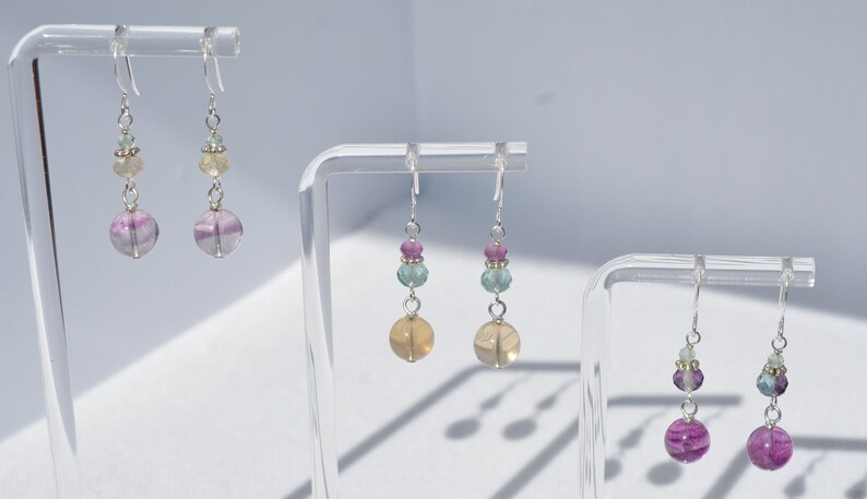 May include: Earrings with a silver hook and a combination of colorful beads. The earrings feature a large, round, purple and white striped bead at the bottom, with smaller beads in shades of pink, green, and yellow above. The earrings are displayed on clear stands.