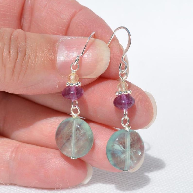 May include: Dangle earrings with silver hooks. Each earring has a large, oval, translucent, blue-green stone, a purple bead, and small, yellow beads. The earrings are held by a hand. The stones are approximately 1.5 cm in diameter.