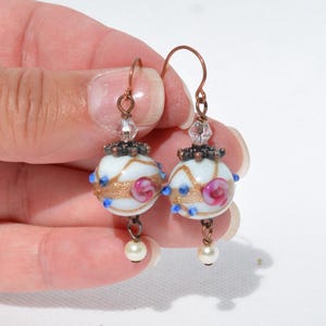 May include: A pair of handcrafted earrings featuring white, round beads with gold and pink floral designs. The beads are accented with blue dots and topped with ornate metalwork and clear crystals. Each earring has a small pearl drop and copper-colored hooks.
