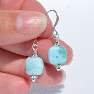 May include: A pair of silver dangle earrings featuring light blue, square-shaped beads. The beads have a slightly rounded shape and are connected to the silver hooks with decorative silver accents. The earrings are displayed against a light background.