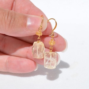Puede incluir: Un par de pendientes colgantes dorados. Cada pendiente presenta un gancho dorado, pequeñas cuentas amarillas y una gema rectangular facetada de color amarillo claro. Los pendientes están sostenidos por una mano sobre un fondo blanco.