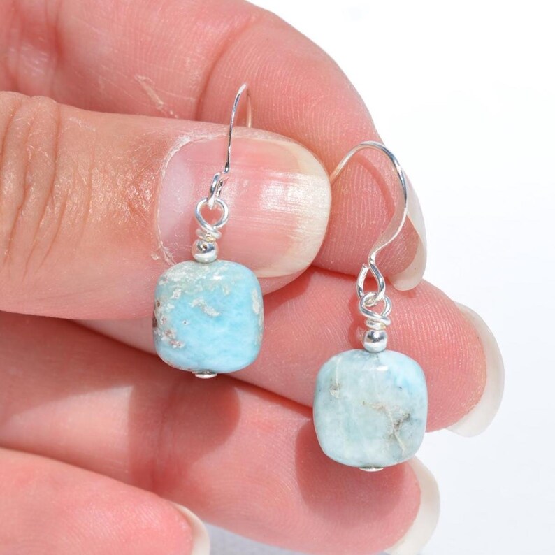 May include: A pair of silver dangle earrings featuring light blue, rectangular gemstones. The gemstones have a marbled appearance with subtle brown and white inclusions. The earrings are held by a hand against a white background.