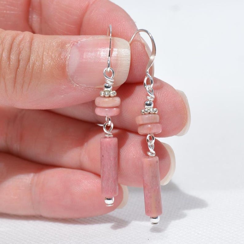 May include: A pair of dangle earrings featuring silver-toned hooks, small silver-toned beads, two flat circular pink stone beads, and one cylindrical pink stone bead at the base. The earrings are constructed with silver-toned wire links.