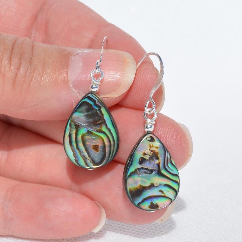 May include: A pair of teardrop-shaped earrings with iridescent abalone shells. The earrings have silver-colored hooks and small silver beads at the top. The abalone shells display a mix of green, blue, and brown colors with a swirling pattern.
