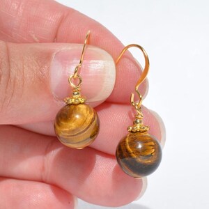 Puede incluir: Un par de pendientes colgantes con piedras preciosas esf&eacute;ricas de ojo de tigre con un patr&oacute;n de bandas marrones y doradas chatoyantes. Cada piedra tiene una peque&ntilde;a tapa de metal dorado y cuelga de un gancho de metal dorado curvado.