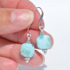 May include: A pair of silver earrings featuring light blue, square-shaped gemstones. The gemstones are connected to silver hooks with decorative silver beads. The earrings are held up by a hand against a white background.