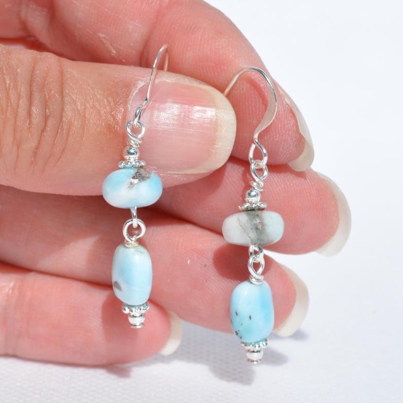 May include: A pair of dangle earrings featuring light blue and white gemstone beads. The earrings have a silver-colored metal hook and decorative accents. The beads are oval and round, with a polished finish. The earrings are displayed against a white background.
