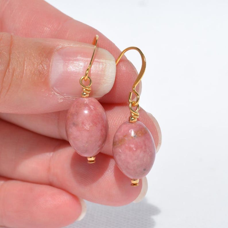 Peut inclure: Une paire de boucles d'oreilles pendantes compos&eacute;es de pierres ovales aux nuances roses et grises marbr&eacute;es, suspendues &agrave; des crochets en m&eacute;tal dor&eacute;. Chaque pierre est fix&eacute;e par un petit fil dor&eacute; enroul&eacute; reliant la gemme au crochet.