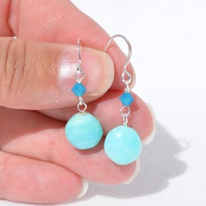 May include: A pair of drop earrings featuring silver-toned hooks, each accented with a small, faceted bright blue crystal bead positioned above a larger, smooth, round, light turquoise blue bead, creating a simple and balanced dangle design.