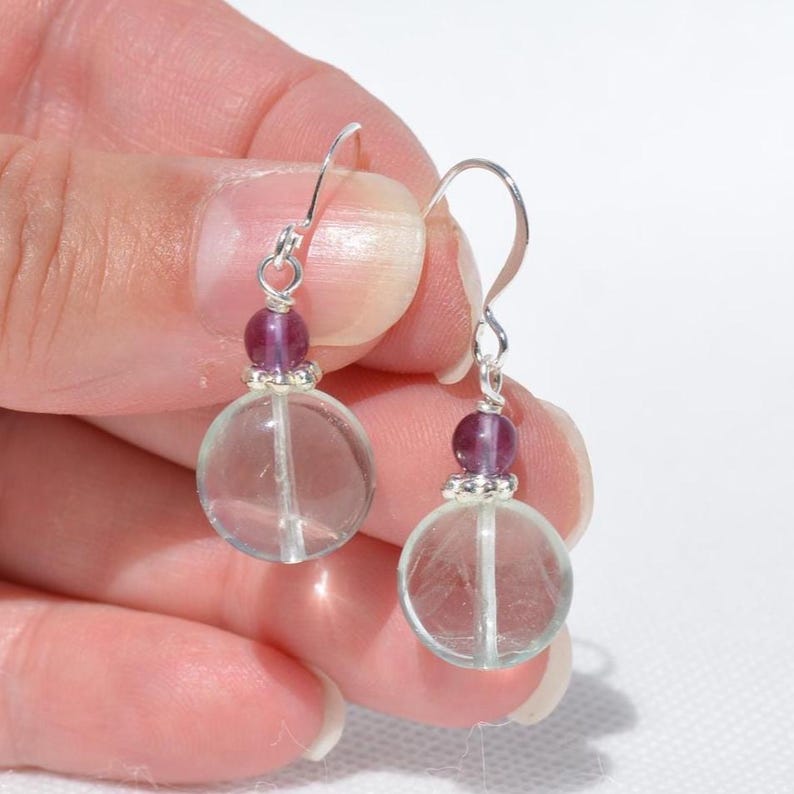 May include: A pair of dangle earrings with clear, round beads and a vertical line, with a small purple bead and silver-tone metal accents. The earrings are held in a hand against a white background.