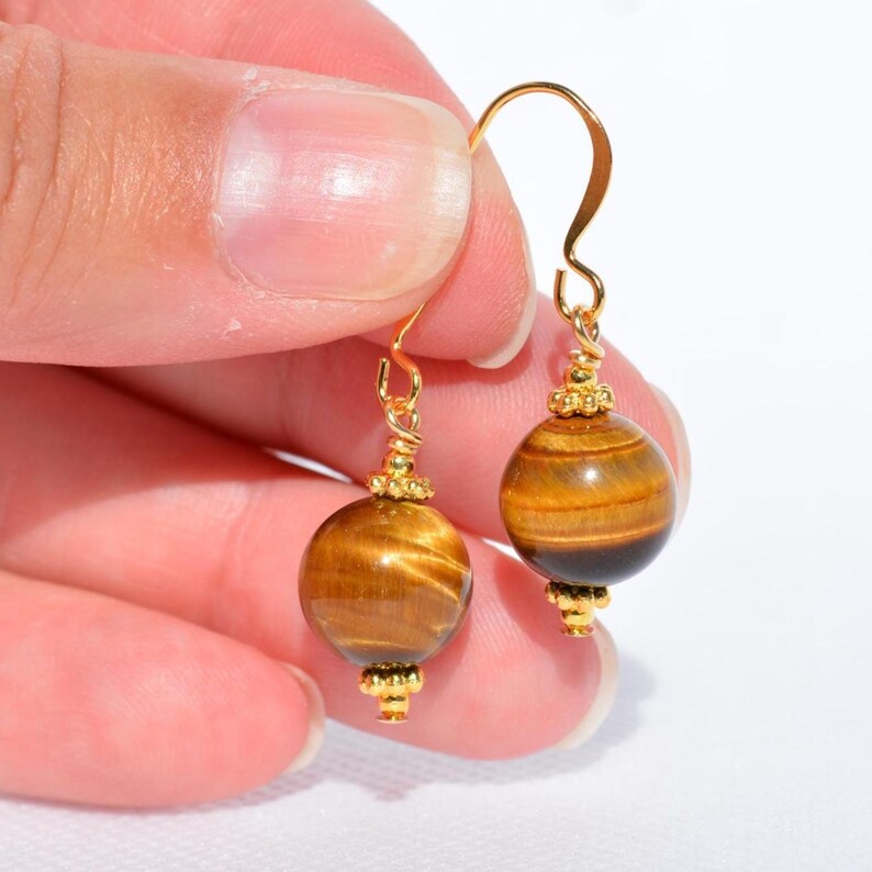 Puede incluir: Un par de pendientes colgantes con cuentas redondas de piedra ojo de tigre con reflejos marr&oacute;n dorado. Cada cuenta est&aacute; decorada con peque&ntilde;as tapas de metal dorado en la parte superior e inferior, suspendidas de ganchos curvos dorados.