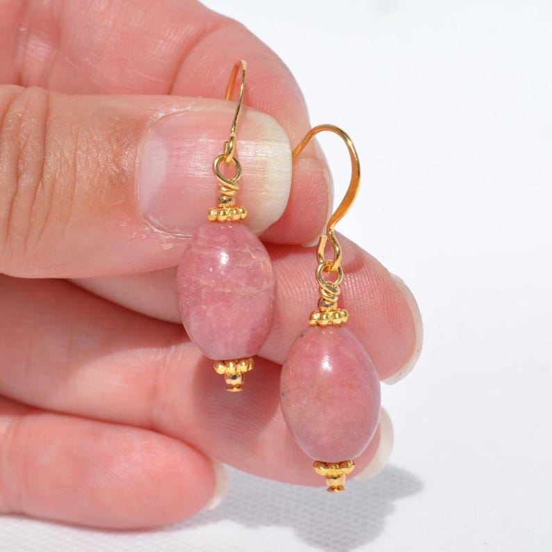 Peut inclure: Une paire de boucles d'oreilles pendantes compos&eacute;es de perles ovales en rhodochrosite rose polie. Chaque perle est agr&eacute;ment&eacute;e de petites perles m&eacute;talliques dor&eacute;es textur&eacute;es en haut et en bas, suspendues &agrave; des crochets d'oreilles dor&eacute;s.