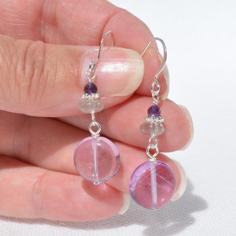 May include: Dangle earrings with a large, round, pale pink gemstone, connected by silver wire to smaller purple and clear beads. The earrings are suspended from silver hooks.