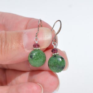 May include: A pair of dangle earrings featuring round green zoisite beads with dark inclusions, accented by small faceted red ruby beads and silver-toned metal spacers, suspended from curved silver-toned ear wires for a simple drop design.