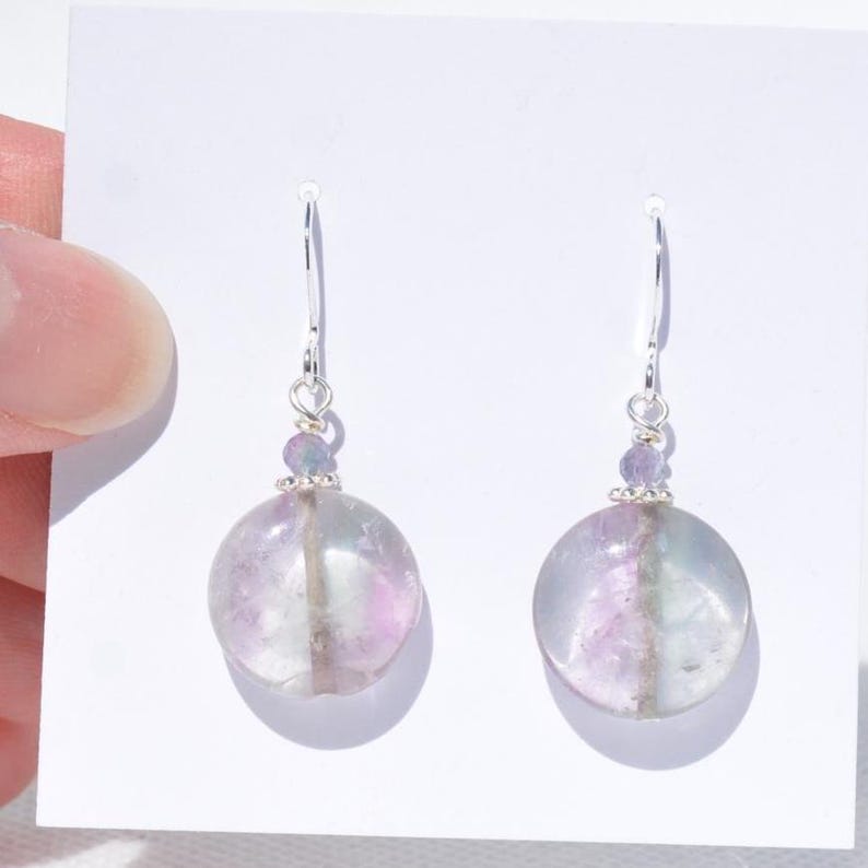May include: A pair of dangle earrings with large, round, translucent fluorite stones in shades of purple, green, and clear. The earrings have silver hooks and small purple beads above the fluorite stones.