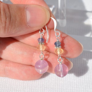 May include: Dangling earrings with a silver hook and wire. Each earring features a large, round, light purple bead at the bottom, with smaller yellow, purple, and silver beads above. The earrings are held in a hand.